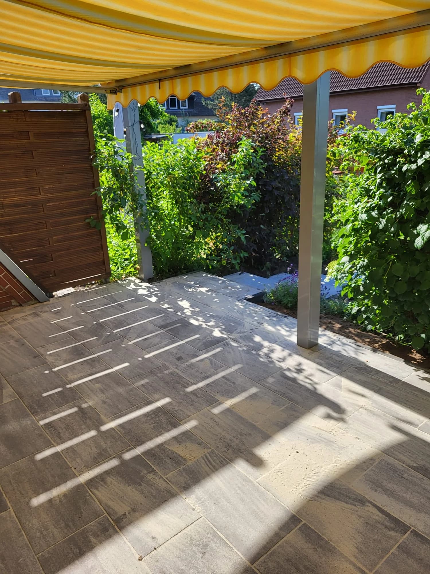 Grau gemusterte Terrassenplatten unter einer gelb gestreiften Markise, dahinter Sträucher